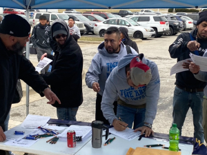 A group of men in sweatshirts gather around a table and sign things, a parking lot is behind them.