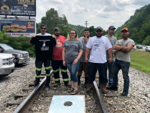 Five people stand on railroad tracks, with a cornhole game at their feet. Some wear "pay us what we are owed" shirts.