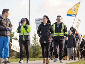 Workers silhouetted against the sky march forward in a line, smiling. Some hold "CUPW" flags aloft. The view is looking up from near the ground, giving them a heroic vibe.