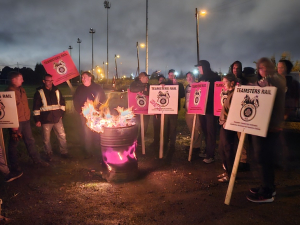 Around dawn, but still dark, a group stand around a burn barrel with signs saying Teamsters Rail locked out