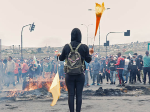 Protester holding what appears to be a flag and a torch in front of burnt out remains. Small crowd in background.