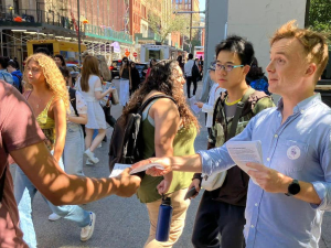 a man in a blue shirt hands a flier to a passerby wearing a backpack