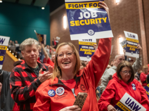 People, mostly wearing red, hoist printed signs in a packed indoor rally. A woman in the foreground, smiling, wears a "no tiers" button. Her sign, held high, reads "UAW: Fight for Job Security." Other visible signs say "UAW: Fight for No Tiers" and "UAW: It's Our Time at Local 2093 American Axle." A man behind her, applauding, wears a red and black buffalo plaid shirt. Most people visible in the photo appear to be white.