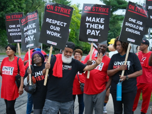 A group of a dozen hotel workers with signs saying “On Strike, Make them Pay” look at the camera, one waves