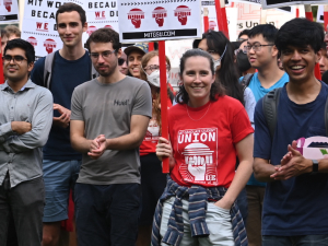 a group of 15 smiles, applauds, and holds signs saying “MIT works because WE do”