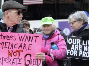 Three older people in a crowd hold signs saying “Our lives are not for Profit” and “Retirees want Medicare, Not Corporate Greed”