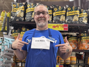 A man in in front of grocery shelves points to a printed-out “Vote No” sign pinned to his blue shop apron.
