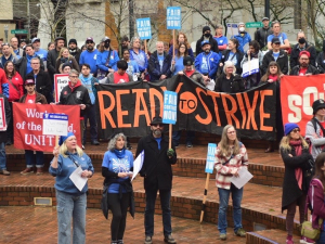 People rally on brick steps (Portland's Pioneer Courthouse Squre) on a rainy day. Many wear blue or red T-shirts under their coats. Banners say "Ready to strike" and "Soldarity." Picket signs say "Fair Contract Now," "Solidarity with Strike-Ready Teachers," and "Essential Wages for Essential Workers."