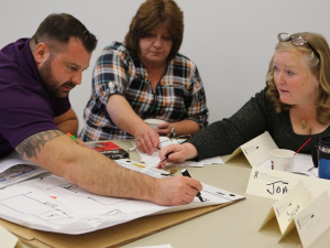 Three people reach across a table to mark a drawing of their workplace