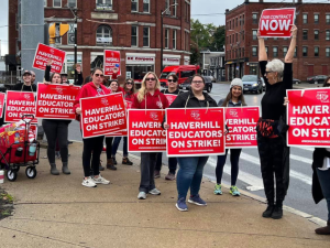 About 15 people, mostly white women, stand on a street corner wearing big red printed signs that say "Haverill educators on strike!" One woman holds her sign high in the air and turns to face the others, in profile to the camera, with a lively open-mouthed expression, maybe yelling or singing. Her sign says "Fair contract now." 