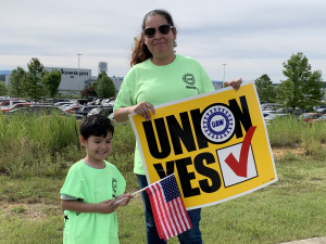 Woman holding "UNION YES" sign and child holding U.S. flag, with VW plant behind