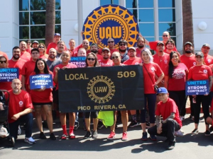 A group of 30 red-shirted autoworkers stand around a banner and hold contract campaign signs.