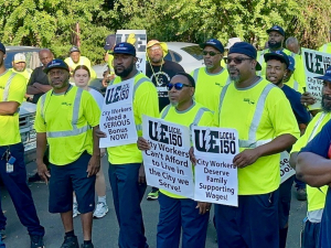 A group of sanitation workers, most in their bright green hi-visibility work vests, hold signs saying, "Fair Pay for City Workers!" and "City Workers Can't Afford to Live in the City we Serve!" during a rally.