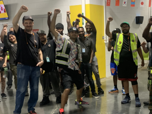 A group of smiling young workers, diverse in race and gender, raise their fists on the warehouse floor. Their pose is casual and triumphant -- looks like they just came from collective action. Some wear safety vests.