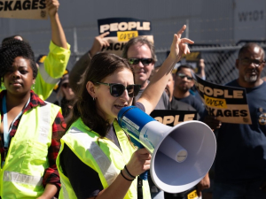 Several workers in green high-viz vests chant and hold signs, one woman in front speaks into a megaphone.