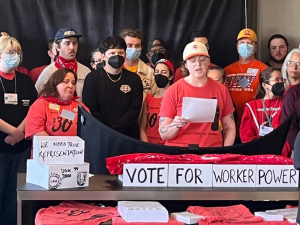 A group of serious-faced people stands indoors. Some are wearing matching red EW4D T-shirts, including Iris Scott, a white woman speaking from notes while others listen and someone in front of them holds up a phone camera to livestream the event. Some in the group wear other union shirts. Handwritten signs on the table in front of the group say "We need true reprsentation," "Vote for worker power," and "UFCW 3000, UAW, TDU" [with hand-drawn logos]. 