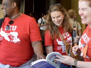 Two women and a man, all wearing Somerville Educators red T-shirts, laugh as they look at a book