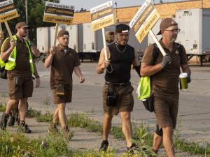 A line of men in brown UPS uniforms and backwards hats with signs saying “Just Practicing for a Just Contract” walk by trailer trucks labeled UPS.