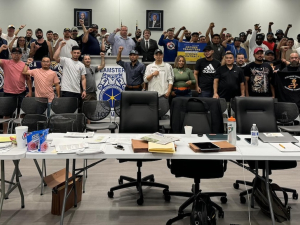 A group of 50 people stand behind a bargaining table with fists raised. 