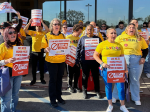 A crowd of people, mostly women, racially varied, in yellow T-shirts (some say UFCW, some say LIBRE) stand outside a grocery store, holding printed red-and-white signs, shouting together. There is a podium. Printed signs have UFCW Local 324 logo and such messages as "Stop the merger," "Say no to higher prices, fewer choices, store closures," and "Protect workers, protect customers, protect community." 