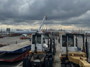 Small port vehicles are in the foreground, and harbor cranes are visible in the distance.