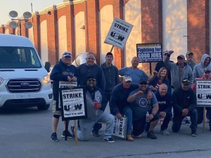 A group of striking steelworkers pose for a photo in a driveway, while a van carrying replacement workers waits behind them.