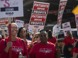 a group of smiling nurses, mostly African American, in red shirts hold signs saying ‘patients before profits’