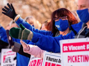 Woman in blue coat facing sideways with hand up among many picketers with hands up, who are holding signs in support of St. Vincent striking nurses