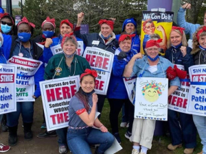 Nurses stand together holding picket signs and wearing red bandanas on their heads, a la Rosie the Riveter.