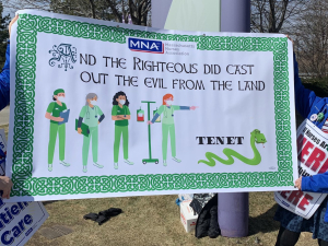 Two nurses hold a green St. Patrick's Day banner that reads "And the righteous did cast out the evil from the land." The banner pictures a group of nurses chasing off a snake labeled Tenet.