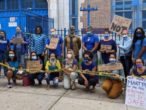 21 adults in masks outside a school. Together they hold "CAUTION" tape. Handmade signs say "100% remote start," "no mas muertes," "ready or not," "we demand safe schools," "no more martyrs for the DOE #Remote until it's safe" and "people got sick in this buidling."