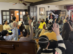 A group of people in chairs crowd a room listening to a speaker.