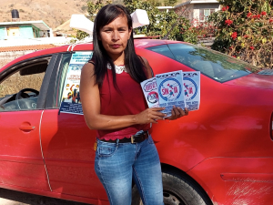 A woman wearing a red blouse and jeans and a serious, determined expression holds up a handful of "VOTA SINTTIA" leaflets, fanned out. She is standing in a sunny outdoor location, in front of a red car that has a poster taped on the side.