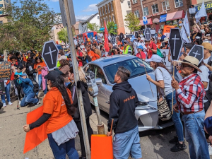 A crowd fills the street holding signs, some of them shaped like coffins.