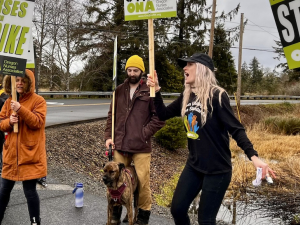 Six people, dressed for a chilly day, holding ONA "Nurses on Strike" picket signs, shout or sing against a gray Oregon sky alongside a road. One man holds the leash of a dog. Most people in the photo appear to be white women.