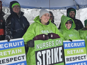 Six smiling people with green and blue strike signs stand under a tent in the snow.