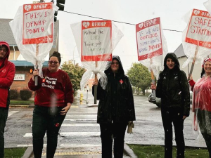 A group of six people stand facing the camera in the rain with signs covered with clear plastic bags. One says, ‘Without the Lab, You’re Just Guessing.’