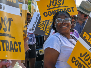 A Black woman in sunglasses smiles at the camera. She is holding a yellow and brown picket sign that says "UPS Teamsters, Just Practicing for a Just Contract." She is surrounded by matching signs, presumably held by other people who are mostly not visible in the shot.