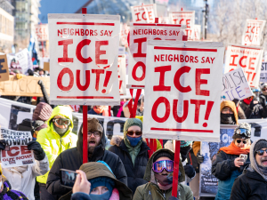 View of a section of a giant crowd marching, on a bright sunny day, bundled against extreme cold. Prominent picket signs that look screen-printed or stenciled say "Neighbors say ICE out!" in crisp red on white background.