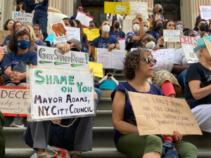 A large crowd of people with handmade signs sit on the steps of a public building. The most visible signs in the foreground say: "Get stuff done! Shame on you Mayor Adams, NY City Council." "The early years are the crucial years. Support our youngest children!" "Reinstate ICs/SWs immediately."