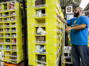 A worker in an Amazon warehouse picks items from a yellow shelf mounted on a robot.