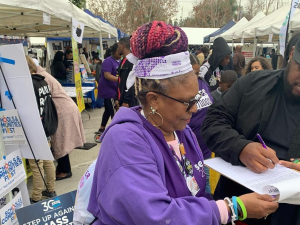 Outdoors at a public event, a woman in SEIU purple holds a clipboard while a man signs. The booth behind her is decked witht "SCHOOLS AND COMMUNITIES FIRST" signs and one that says "Step up against mass incarceration."