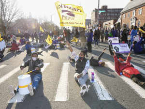 Several women sit in the middle of the crosswalk, holding upside-down buckets that they are using as drums. One holds a flag that reads "SEIU 1199 New England: Health Care for All."