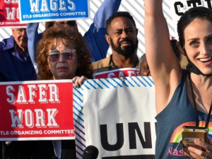 A group of mostly women stand close together with signs saying “Higher Wages” and “Safer Work”