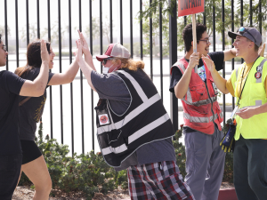 A woman in a safety vest high-fives two others in front of a fence while others stand by with strike signs