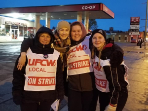 Four smiling women wearing "UFCW Local ON STRIKE" signs stand in front of a Co-op gas station, arms around each other's shoulders