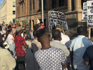 A group of workers, mostly Black, face an official-looking building with signs.