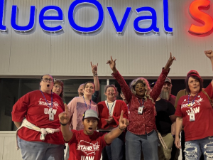 A group of 7 workers look excited and raise fists in front of a “BlueOval SK” sign