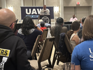 An audience listens to a man at a lectern. One audience member has a blue shirt with “Union Strong” on the back.