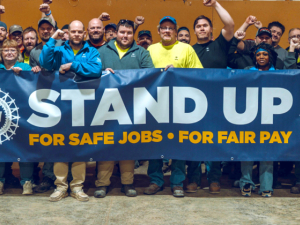 Thirty people, many with fists raised stand behind a banner that says “Stand Up, for safe jobs, for fair pay”